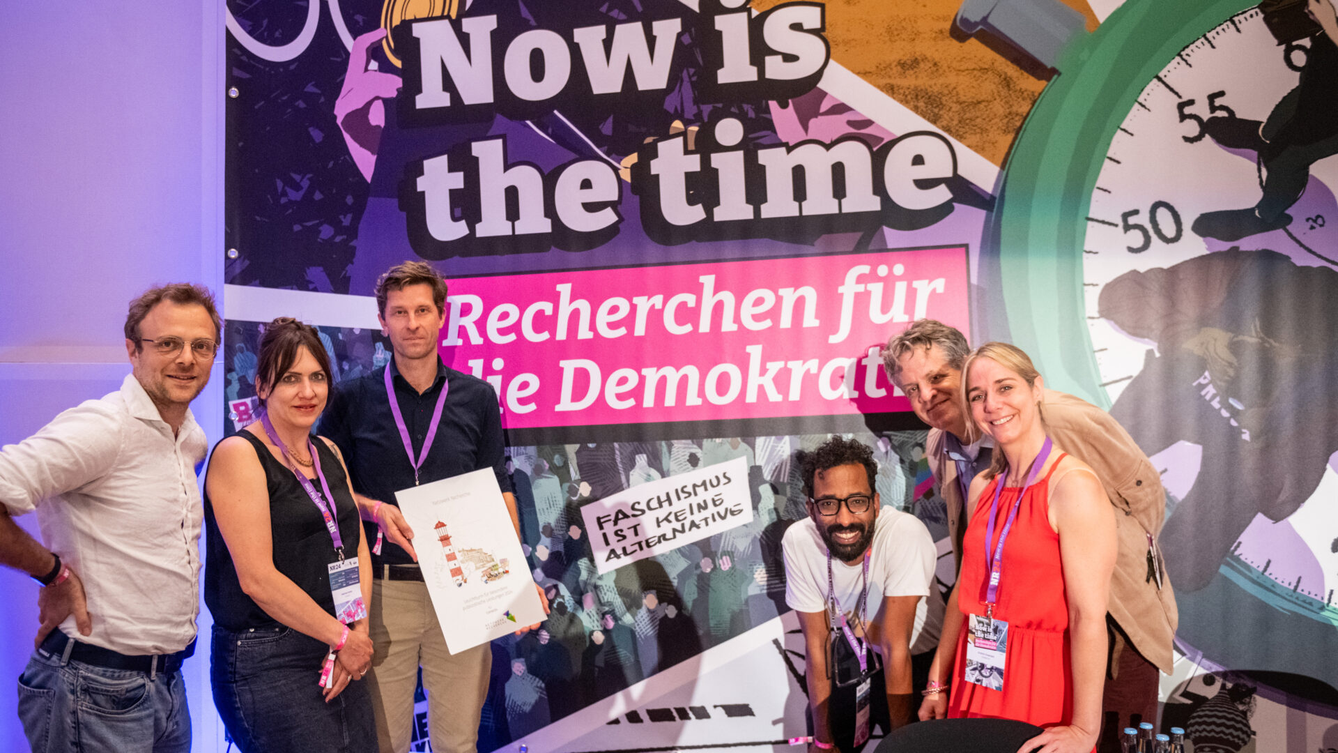 Mehrere Menschen, grinsend in die Kamera. Jemand hält eine Urkunde in der Hand. Im Hintergrund sieht man eine große Leinwand mit "Now Is The Time – Recherchen für die Demokratie".