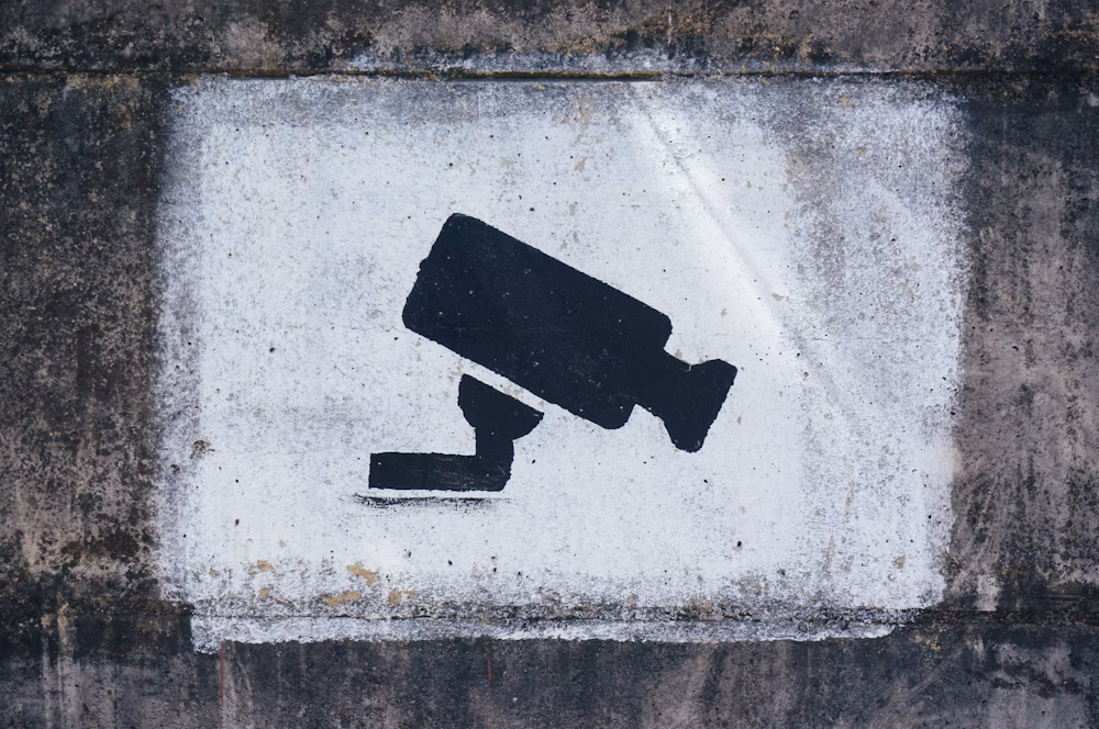 Ein Grafitti-Stencil, gesprayt an eine Betonwand. Es ist eine Videoüberwachungskamera auf weißem Hintergrund zu sehen.